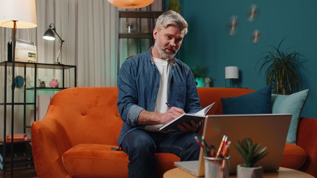 Middle-aged man freelancer teacher study with notebook sitting on sofa at home. Mature guy conference video call talking to webcam at night room. Online e-learning in remote chat. Distance education