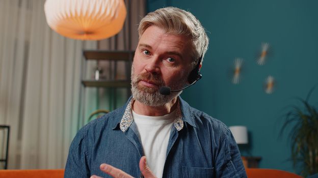 POV of senior man at home wearing headset, freelance worker, call center or support service operator helpline, having talk with client or colleague communication support. Mature guy in evening room