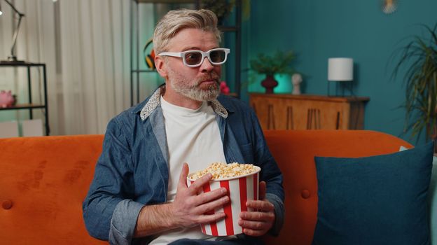 Excited senior mature man sits on couch eating popcorn snacks and watching interesting TV serial sport game, film, online social media movie content at home. Elderly guy enjoying evening entertainment