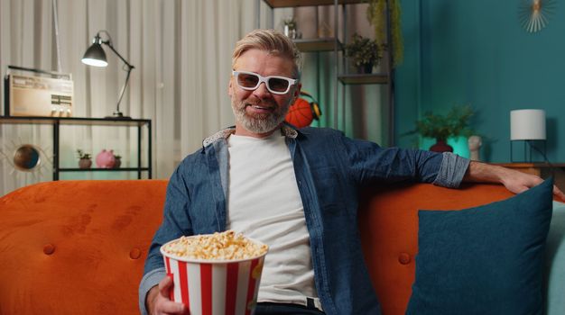 Excited bearded middle-aged man sits on sofa eating popcorn snacks and watching interesting TV serial, sport game, film, online social media movie content at home. Guy enjoying evening entertainment