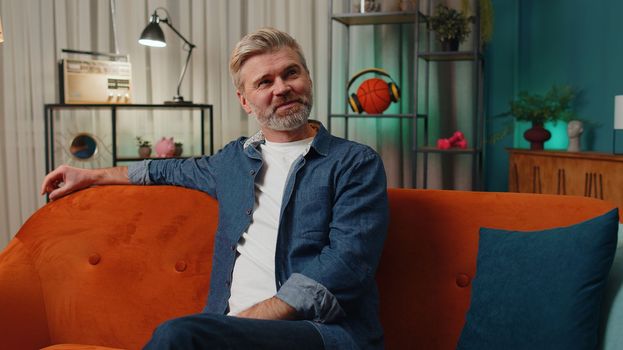 Happy calm middle-aged senior man at home couch smiling friendly, glad expression looking away dreaming resting, relaxation feel satisfied concept good news, celebrate win. Mature guy in night room