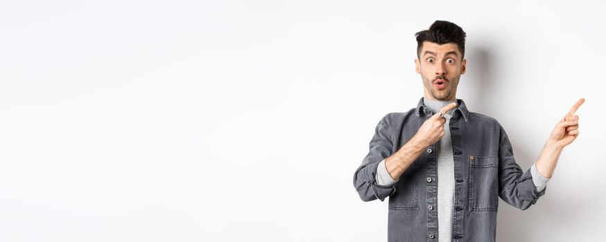 Excited guy showing advertisement aside, pointing fingers right and gasping amazed, checking out special deal, standing on white background.