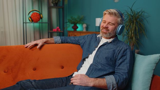 Happy overjoyed middle-aged bearded man in wireless headphones relaxing sitting on couch at home choosing listening favorite energetic disco dancing music. People weekend evening leisure activities