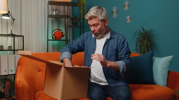 Happy surprised winner man unpacking delivery parcel at home. Smiling satisfied mature guy shopper online shop customer opening cardboard box receiving purchase gift headphones by fast postal shipping