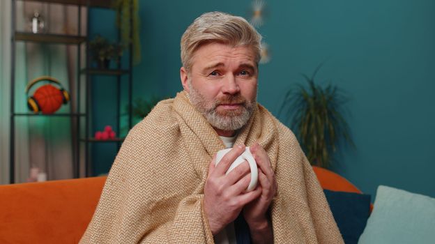 Sick man wrapped in plaid sit alone shivering from cold on sofa drinking hot tea in unheated apartment without heating due debt. Unhealthy mature guy feeling discomfort try to warm up at evening home