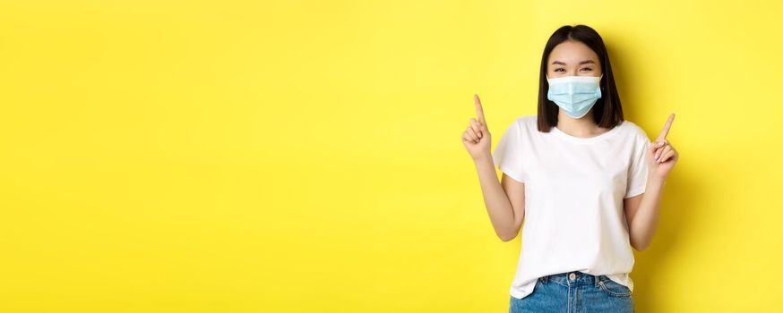 Covid-19, pandemic and social distancing concept. Young asian woman in white t-shirt and medical mask from coronavirus, pointing fingers up, showing special offer.