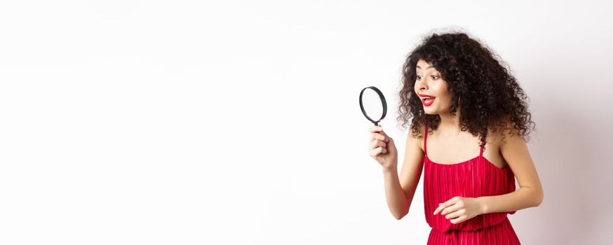 Beautiful woman in red dress and makeup looking at something with magnifying glass, checking out interesting promo, standing over white background.