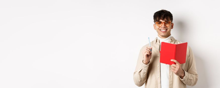 Happy person in glasses writing in journal, holding pen and diary, smiling at camera, plan a schedule, standing with planner on white background.