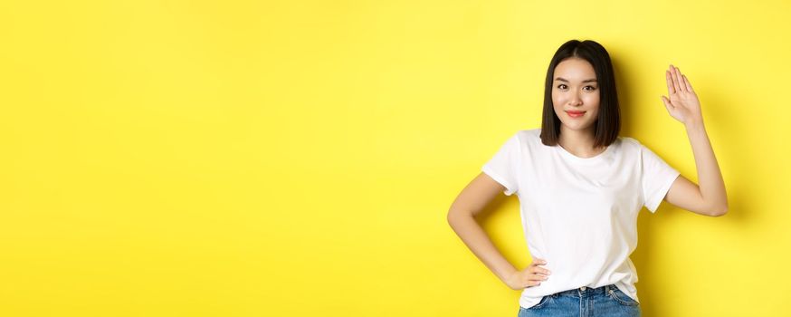 Friendly asian woman in white t-shirt waving hand and saying hello, greeting you, standing over yellow background.