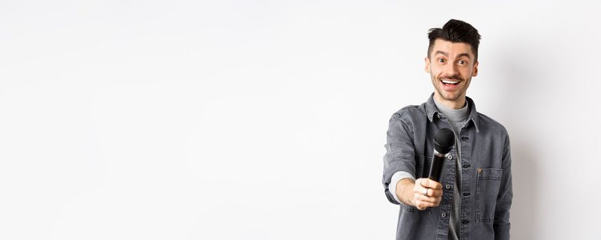 Handsome smiling man stretch out hand with microphone, giving you mic, standing on white background.