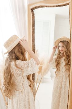 A beautiful teenage girl with long hair measures a straw hat in front of a mirror. Self-admiration of a blonde. selective focus