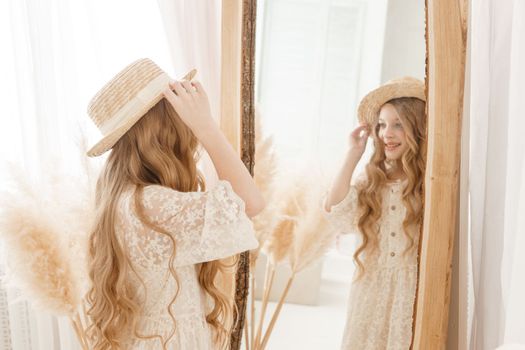 A beautiful teenage girl with long hair measures a straw hat in front of a mirror. Self-admiration of a blonde. selective focus