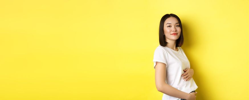 Asian woman smiling kindly, touching belly and looking at camera, being pregnant, having a baby, standing over yellow background.