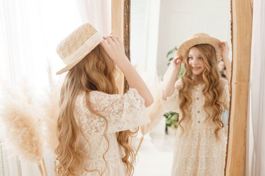 A beautiful teenage girl with long hair measures a straw hat in front of a mirror. Self-admiration of a blonde. selective focus
