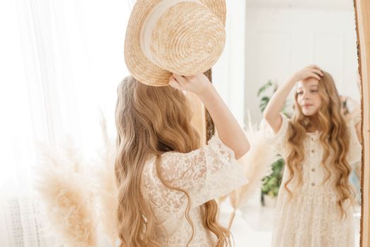 A beautiful teenage girl with long hair measures a straw hat in front of a mirror. Self-admiration of a blonde. selective focus