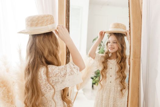 A beautiful teenage girl with long hair measures a straw hat in front of a mirror. Self-admiration of a blonde. selective focus