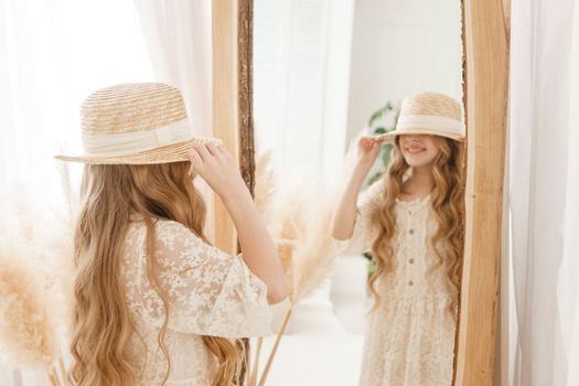 A beautiful teenage girl with long hair measures a straw hat in front of a mirror. Self-admiration of a blonde. selective focus