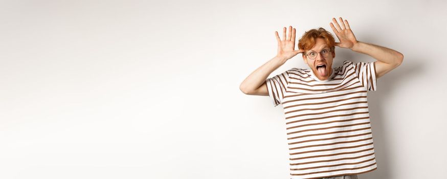 Funny young redhead man acting silly, showing tongue and mocking someone with stupid face, standing over white background.