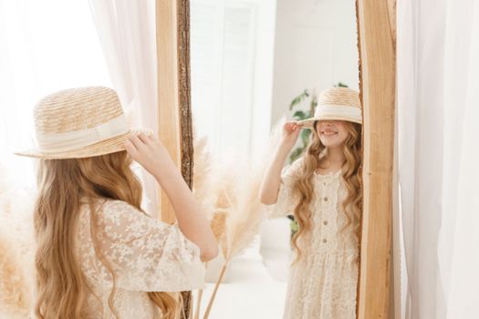 A beautiful teenage girl with long hair measures a straw hat in front of a mirror. Self-admiration of a blonde. selective focus