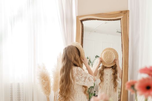 A beautiful teenage girl with long hair measures a straw hat in front of a mirror. Self-admiration of a blonde. selective focus