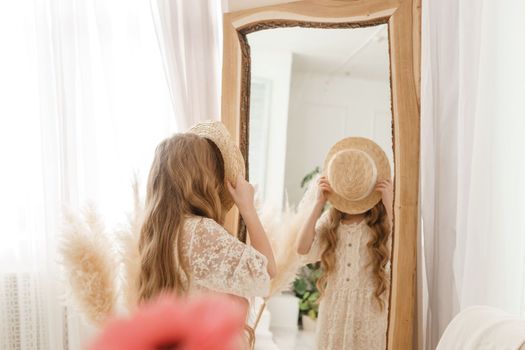 A beautiful teenage girl with long hair measures a straw hat in front of a mirror. Self-admiration of a blonde. selective focus