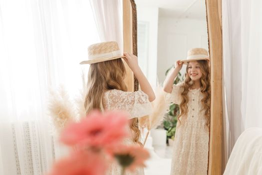 A beautiful teenage girl with long hair measures a straw hat in front of a mirror. Self-admiration of a blonde. selective focus