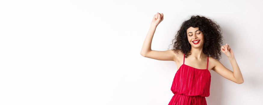 Carefree woman with curly hair, dancing at party, wearing red dress, relaxing with music, standing on white background.