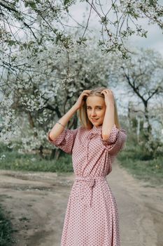 Blonde girl on a spring walk in the garden with cherry blossoms. Female portrait, close-up. A girl in a pink polka dot dress