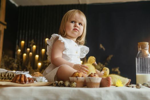 A little girl is sitting on the Easter table and playing with cute fluffy ducklings. The concept of celebrating happy Easter