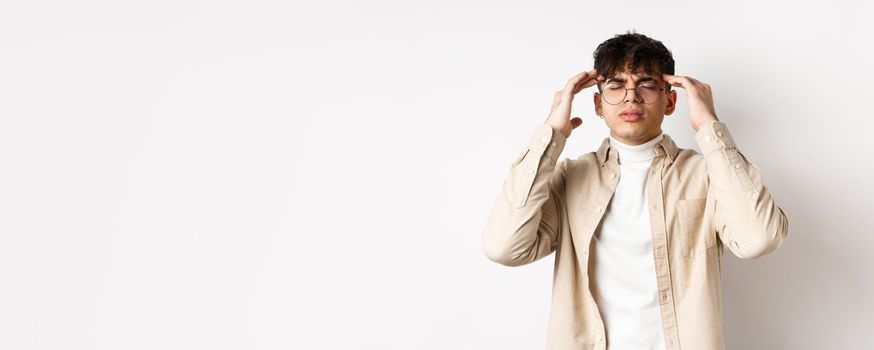 Image of young man in glasses having headache, touching head temples and frowning from painful migraine, feel dizzy, standing on white background.