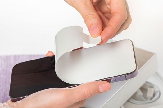 Unpacking a new phone. Remove the protective white film from the new phone. Close-up of a woman's hands removing a protective film from a phone in a purple case. Copy space on white background
