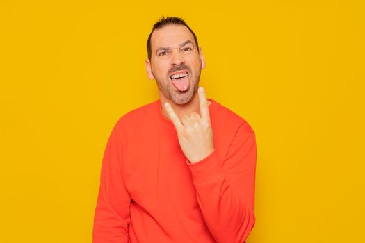 Bearded hispanic man wearing red pullover isolated over yellow background showing rock gesture with fingers and sticking out tongue