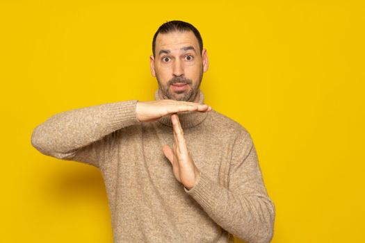 Bearded hispanic man wearing a turtleneck sweater showing time out gesture, tired of a lot of work. Indoor studio shot isolated on yellow background