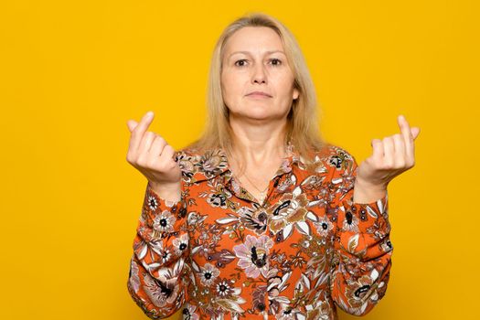 Pretty caucasian blonde woman in a patterned dress making the money gesture with her fingers isolated over yellow background