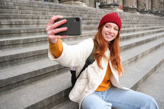 Young redhead tourist takes selfie in front of museum on stairs, holds smartphone and looks at mobile camera, makes photo of herself with phone.