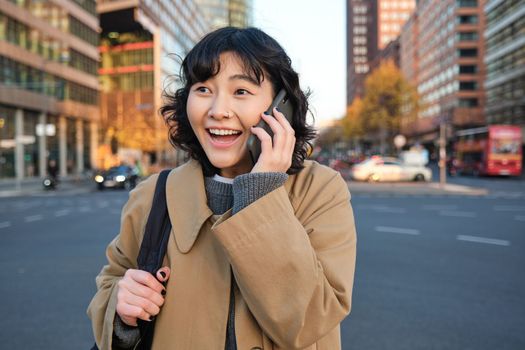 Portrait of happy korean girl, talks on mobile phone, looks surprised and happy, receives positive great news over telephone conversation, stands on street of city.