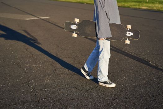 Cropped shot of teen girl body, holding cruiser longboard in hand, walking in sneakers on road in jeans and sweatshirt. Young woman skater with skateboard.