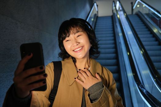 Stylish hipster girl in headphones, going down escalator and video chats, records herself on smartphone, looks at mobile phone camera.