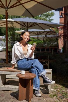 People and communication. Stylish asian girl sits in outdoor cafe with cup of coffee and smartphone, using mobile phone.