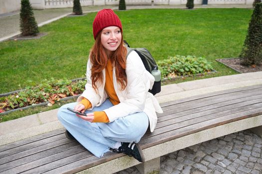 Portrait of stylish young woman, 25 years, sits on bench in park and uses mobile phone, reads online news, messages or watches video on smartphone app, connects to public wifi.