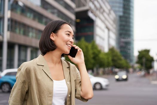 Portrait of smiling asian woman talking on mobile phone, walking on street near busy road and speaking to friend, laughing.