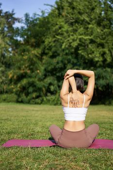 Rear view of sportswoman in middle of workout, stretching her amrs behind back, sitting on rubber yoga mat on lawn in park.