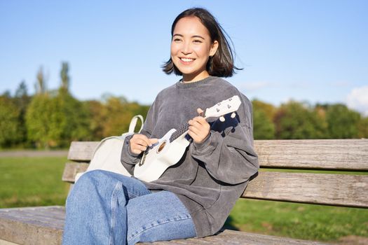 Happy cute girl sits alone on bench in park, plays ukulele guitar and enjoys sunny day outdoors.