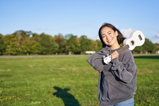 Portrait of beautiful smiling girl with ukulele, asian woman with musical instrument posing outdoors in green park. People and lifestyle