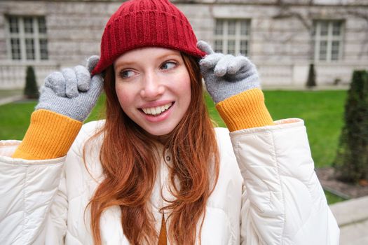 Close up portrait of beautiful redhead woman in red knitted hat, warm gloves, smiling and looking happy at camera, sitting in park.