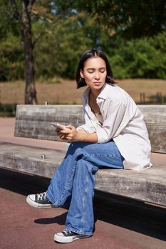 Portrait of asian girl sitting with smartphone feeling sad, looking gloomy and frustrated, waiting for a call outdoors in park.
