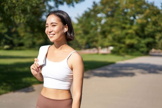 Smiling asian fitness girl holding towel on shoulder, workout in park, sweating after training exercises outdoors.