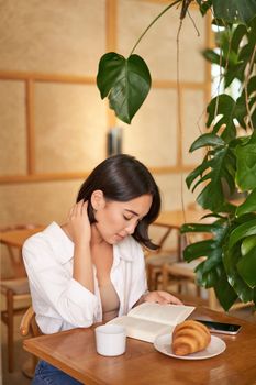 Beautiful young asian woman sitting in cafe with a book, eating croissant and reading, drinking cup of coffee. Wellbeing and self-care