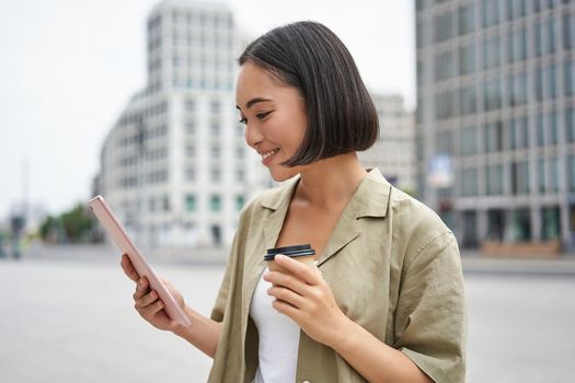 Portrait of smiling korean girl looks at tablet, reads on street, drinks takeaway coffee.