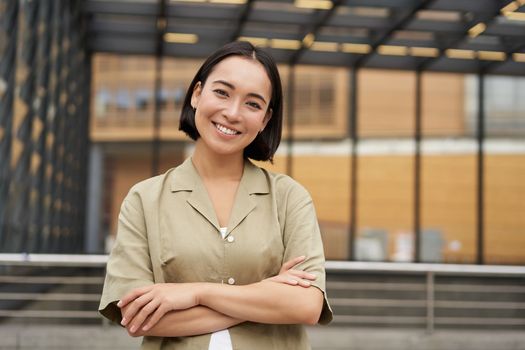 Portrait of young asian woman standing with confidence, cross arms on chest and smiling, posing outdoors on street.